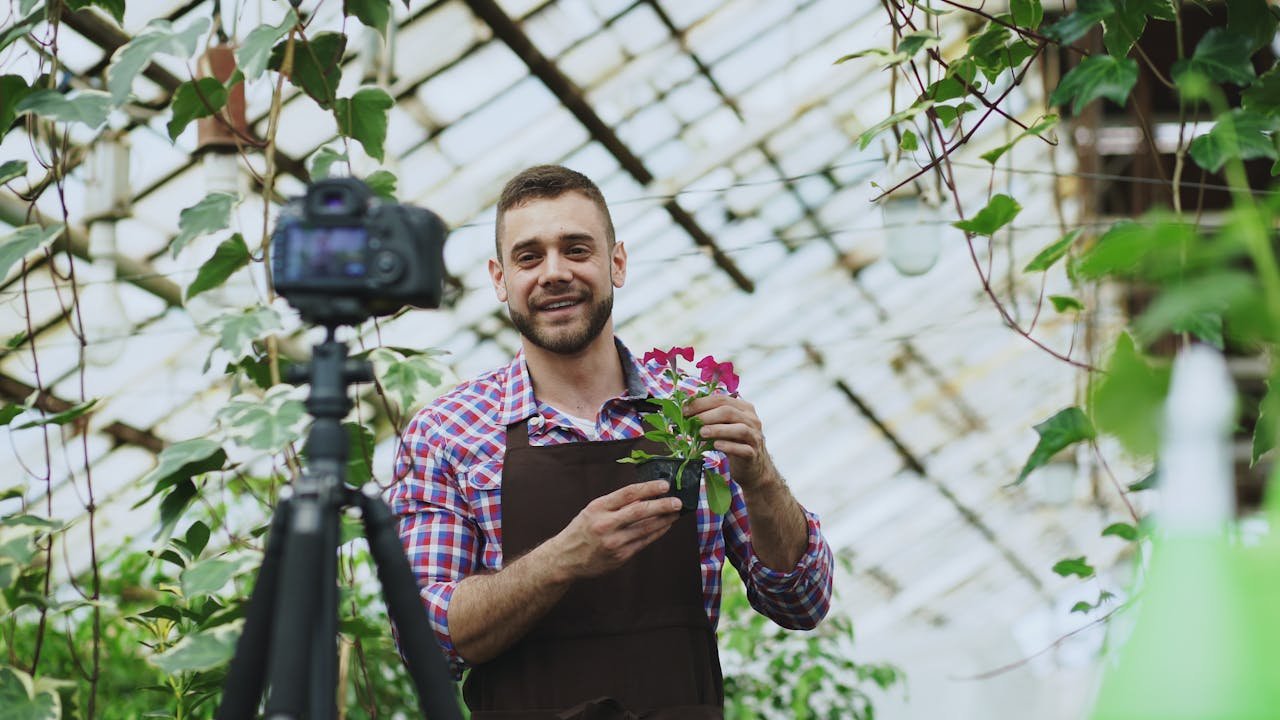 A gardener in a greenhouse filming a tutorial with a digital camera, holding flowers.