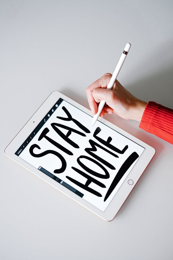 A hand writes 'Stay Home' on an iPad using an Apple Pencil, highlighting remote work and technology.